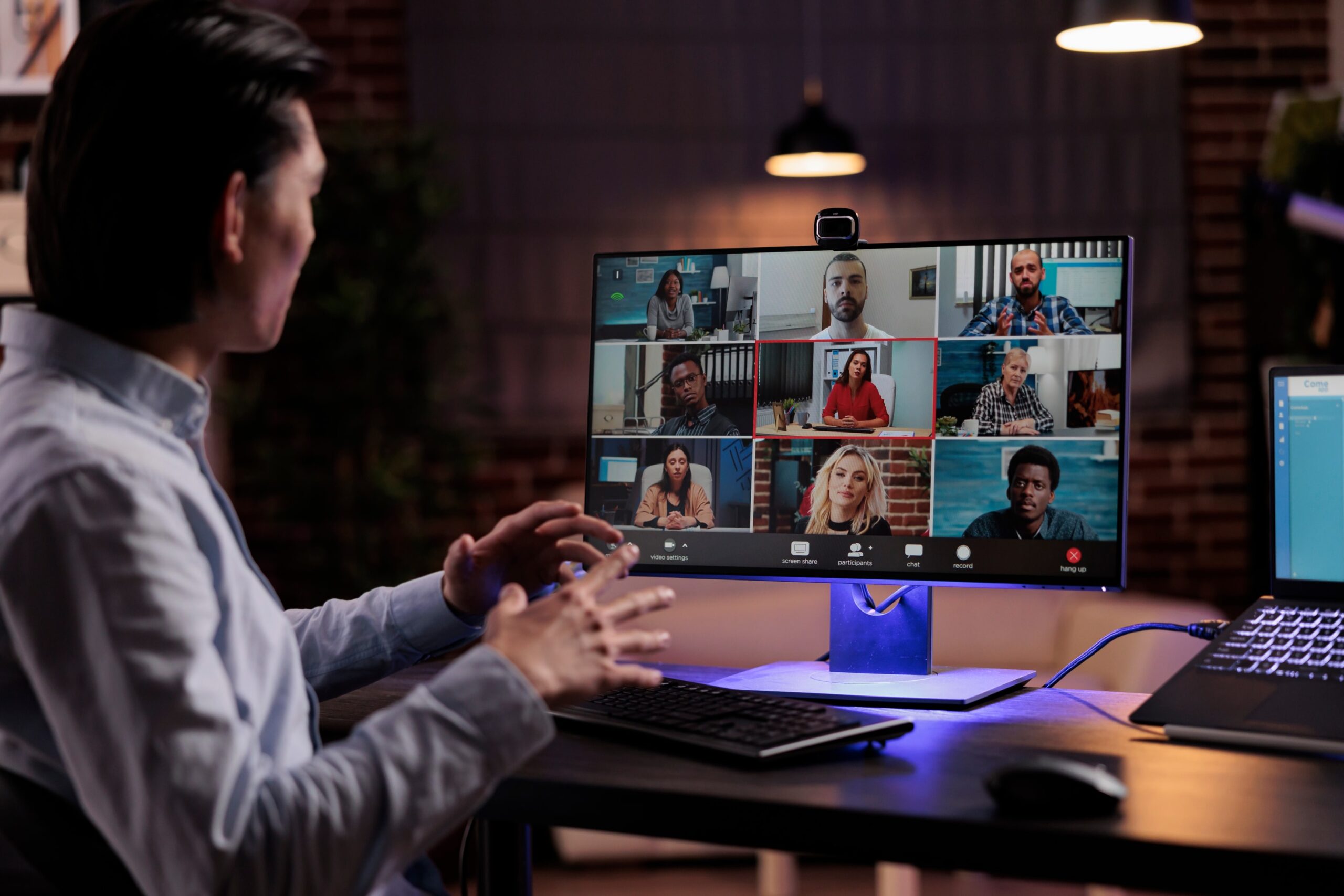 A man at a desk uses a laptop while engaging with multiple participants displayed on a computer screen.